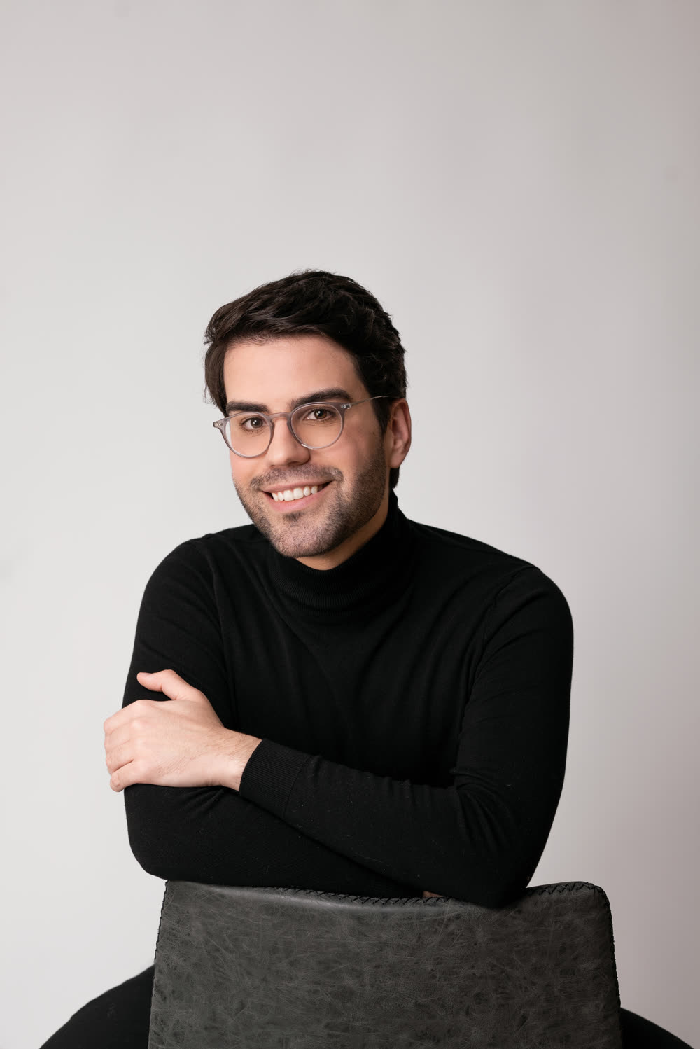 Lucas Bulcão sorrindo, de blusa preta de gola alta, apoiado numa cadeira de madeira